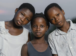 Three children standing shoulder-to-shoulder outdoors.
